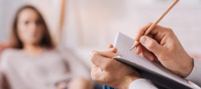 Therapist taking notes during psychotherapy session with patient in a clinical setting.
