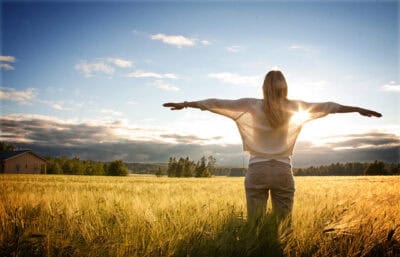 Woman standing in a field with arms outstretched at sunset, symbolizing wellness and serenity.