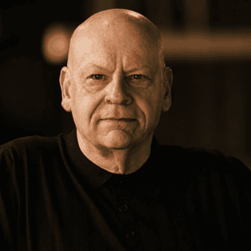 Close-up of a mature man in a black shirt, looking serious, in a professional setting.
